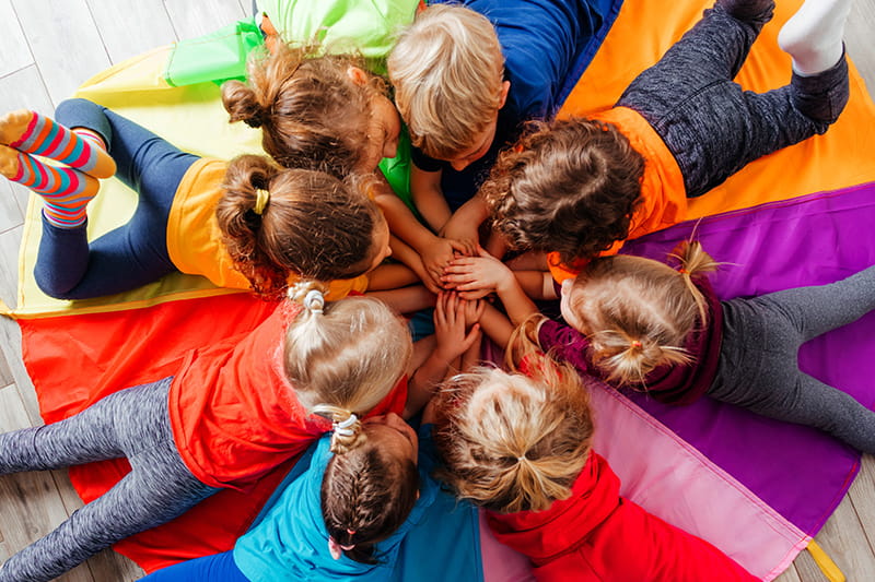  Acht Kinder liegen auf einem Regenbogentuch und haben die Hände als Team zusammengefaltet.