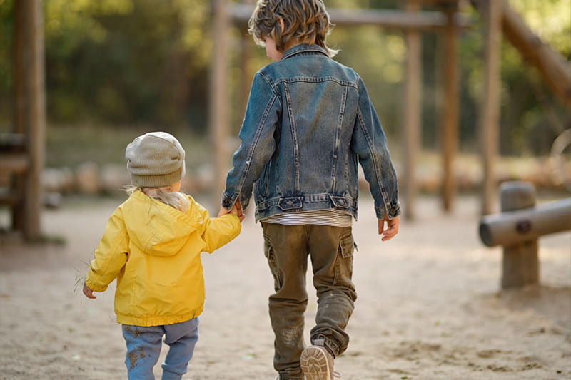 Zwei Kinder unterschiedlichen Alters laufen Hand in Hand über einen Spielplatz.